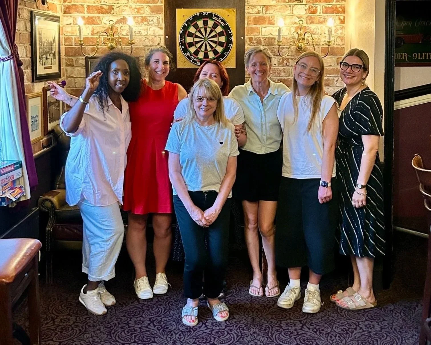 Group of eight women standing in a pub with brick walls and a dartboard in the background.