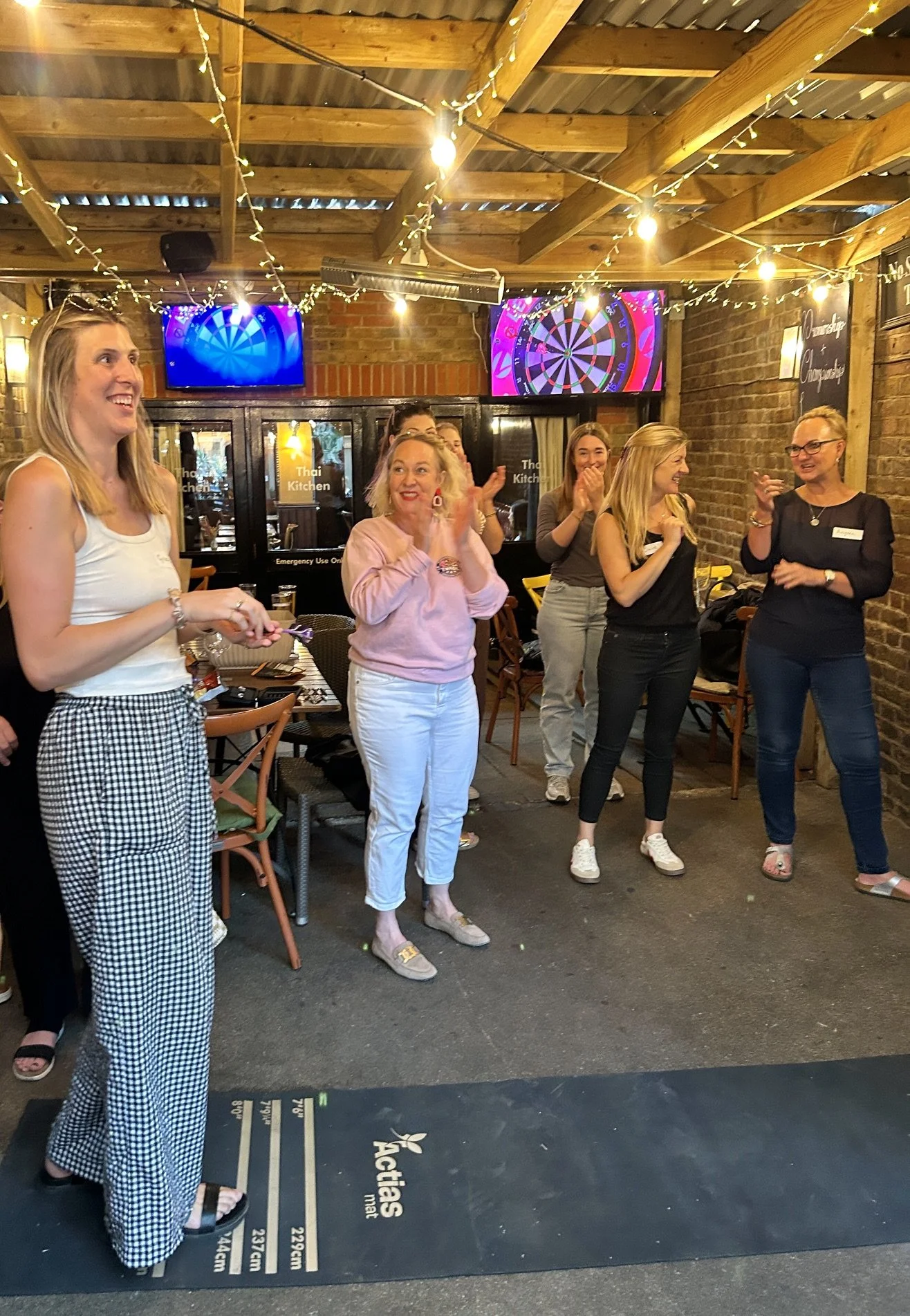 Group of women celebrating or socializing in a rustic restaurant or bar with wooden beams, string lights, and dartboards on the wall, some clapping and smiling.