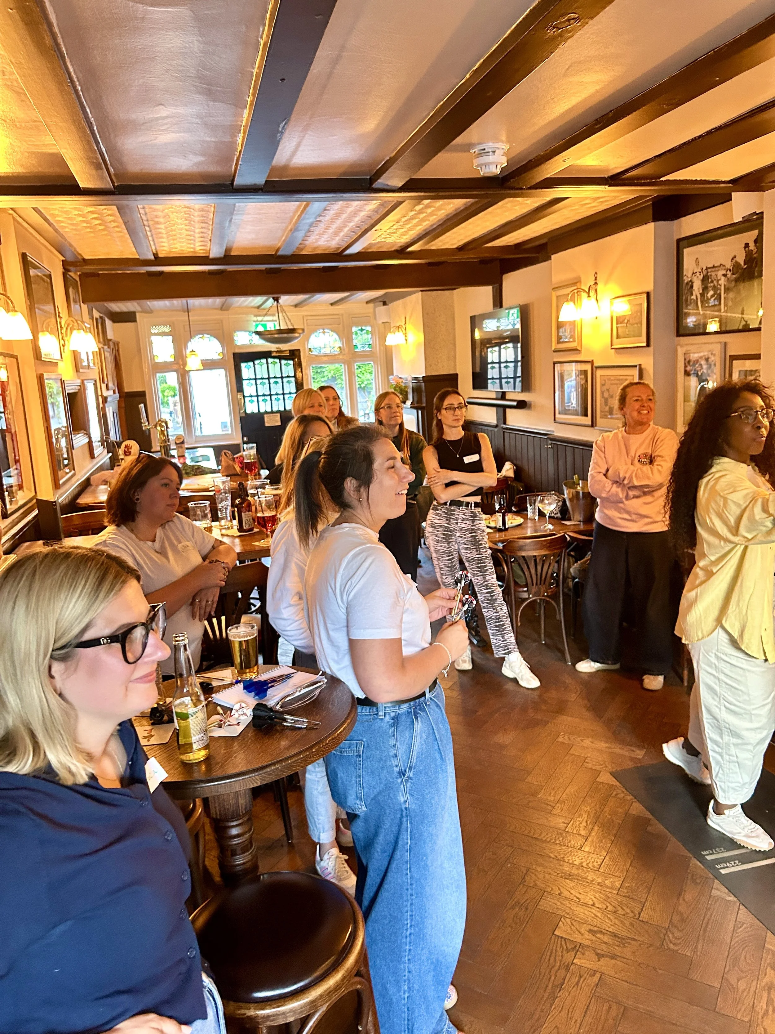 A group of women in a cozy restaurant or pub, some are sitting at a round table with drinks, while others are standing and listening, with warm lighting, wooden ceiling beams, and framed pictures on the wall.