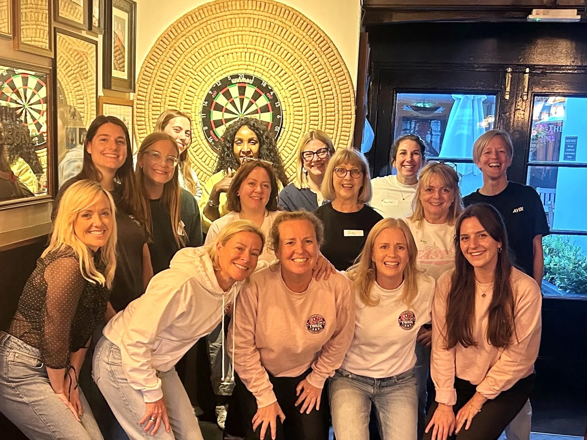 Group of women smiling and posing for a photo in a bar or pub with a dartboard on the wall behind them.