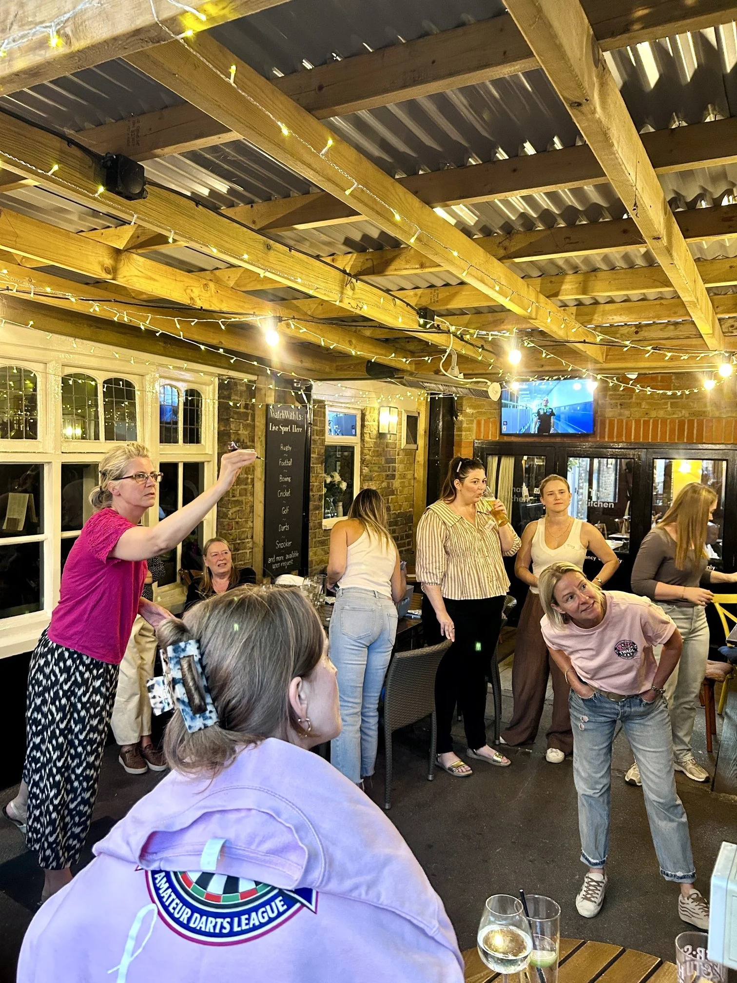 A group of women gathered inside a pub or restaurant, some standing and some seated, with a woman in a pink shirt and black and white patterned pants gesturing with her right hand. There are drinks on the table, string lights on the ceiling, and a te
