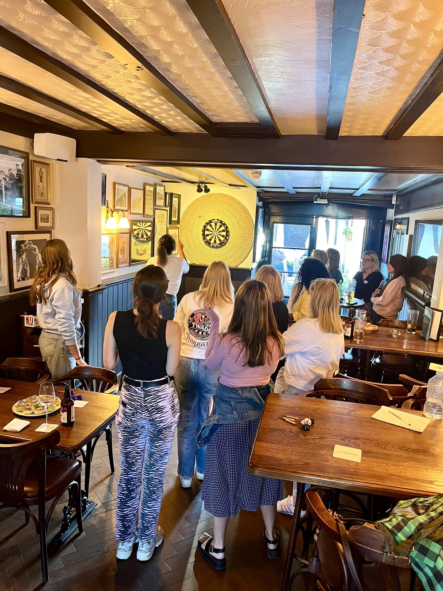 A group of women gathered in a pub or bar, watching darts on a TV screen. Some are standing, while others are sitting at tables. The room is decorated with framed pictures and has a dartboard on the wall.