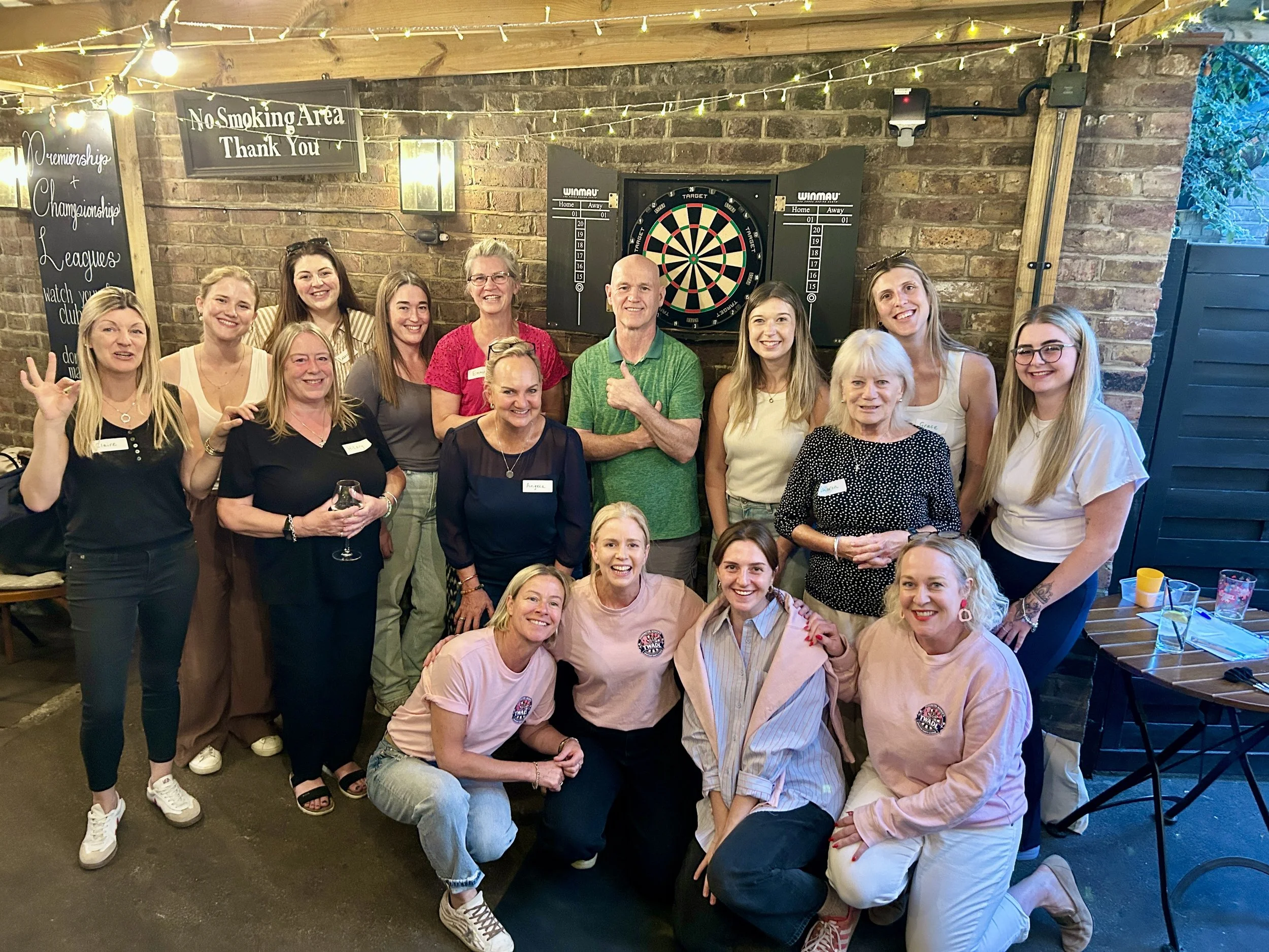 A group of people, mostly women, posing for a photo in a rustic indoor space with a brick wall background, a dartboard, and string lights hanging overhead. Many are smiling and some are holding drinks.