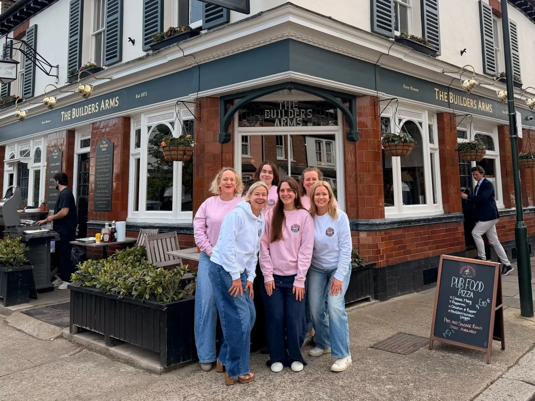A group of six women standing outside a pub called The Builders Arms, smiling at the camera. The pub has a blue and white exterior with hanging flower baskets and large windows. Some people are preparing food or standing near the entrance.