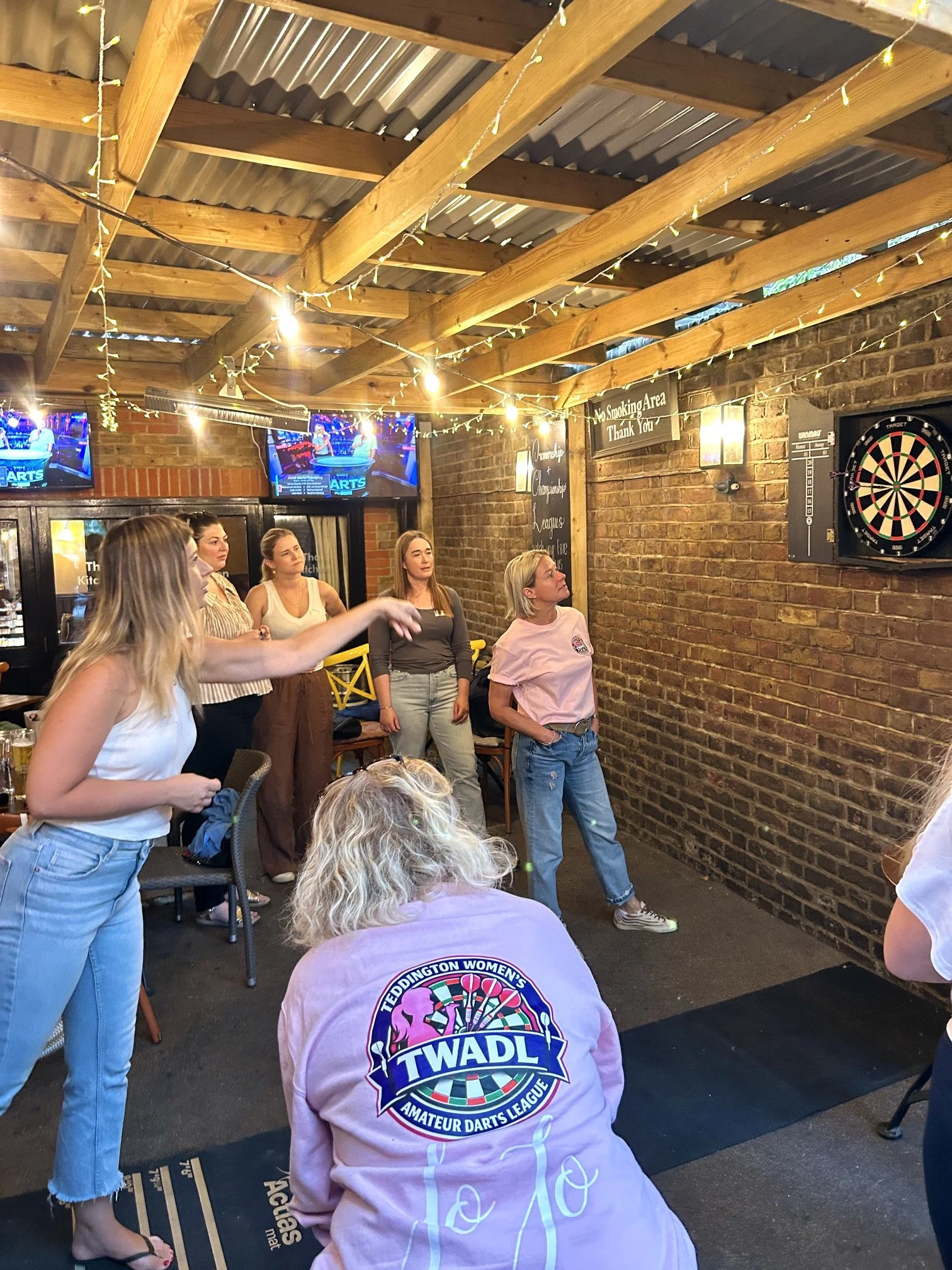 People playing darts in a dimly lit bar or game room with brick walls and string lights overhead.