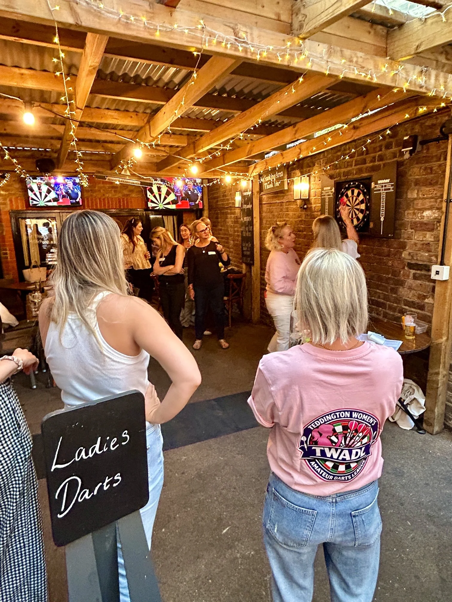 A group of women gather inside a rustic bar or darts lounge, decorated with string lights and brick walls, with women talking and socializing.