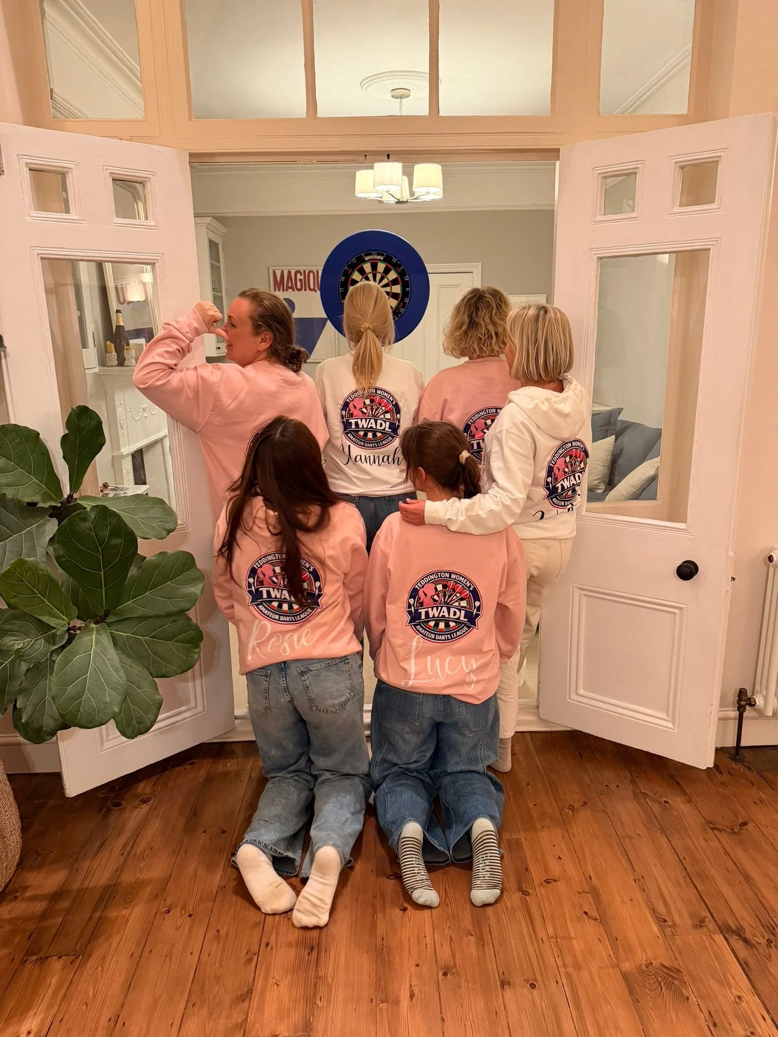 Group of women in matching pink and white sweatshirts with logo, kneeling and standing in front of a dartboard, inside a cozy home with wooden floors and a potted plant in the foreground.