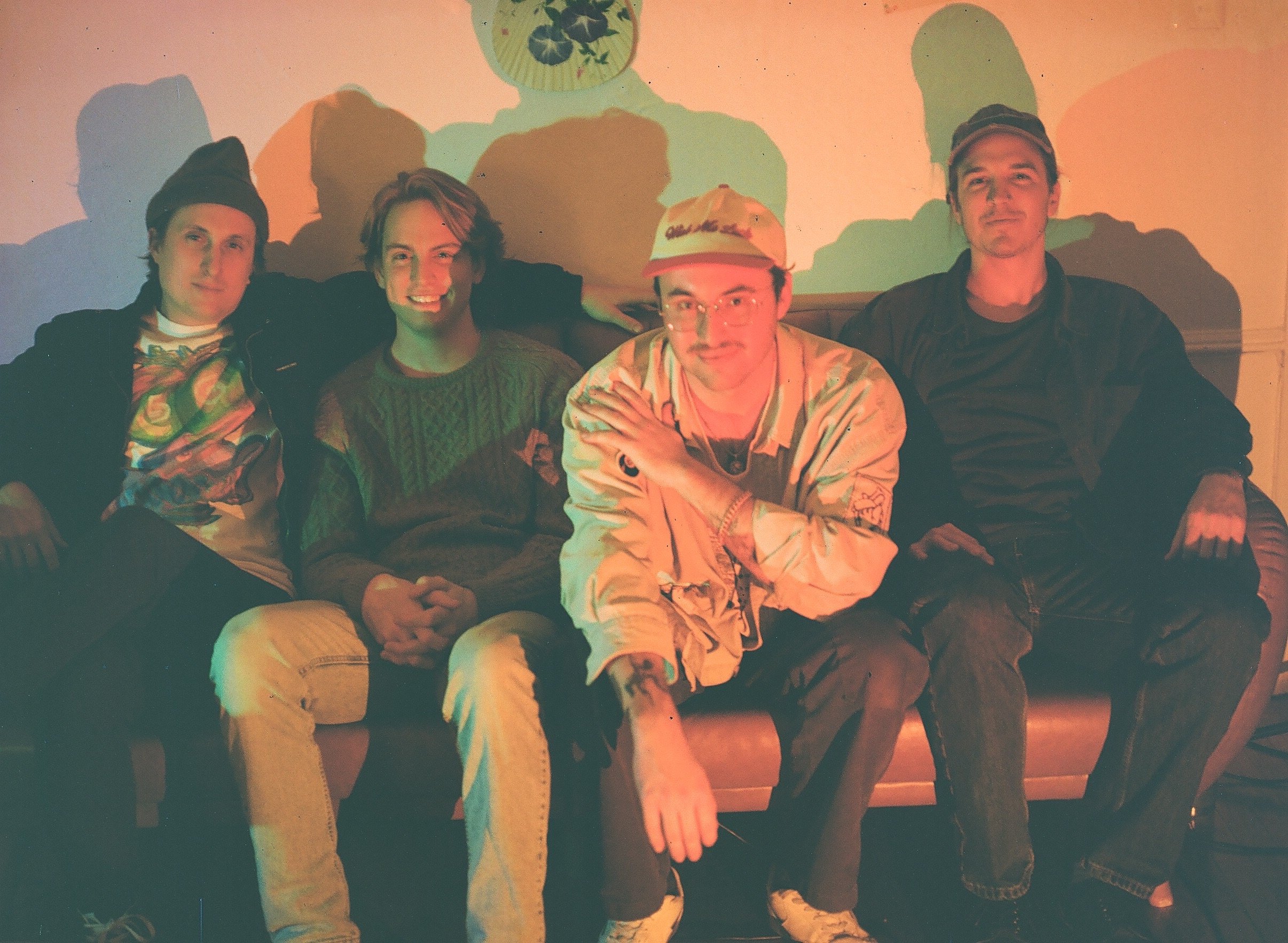 Four young men sitting on a couch against a colorful wall with shadows cast behind them. They are casually dressed, with some wearing hats, and are smiling or looking relaxed.