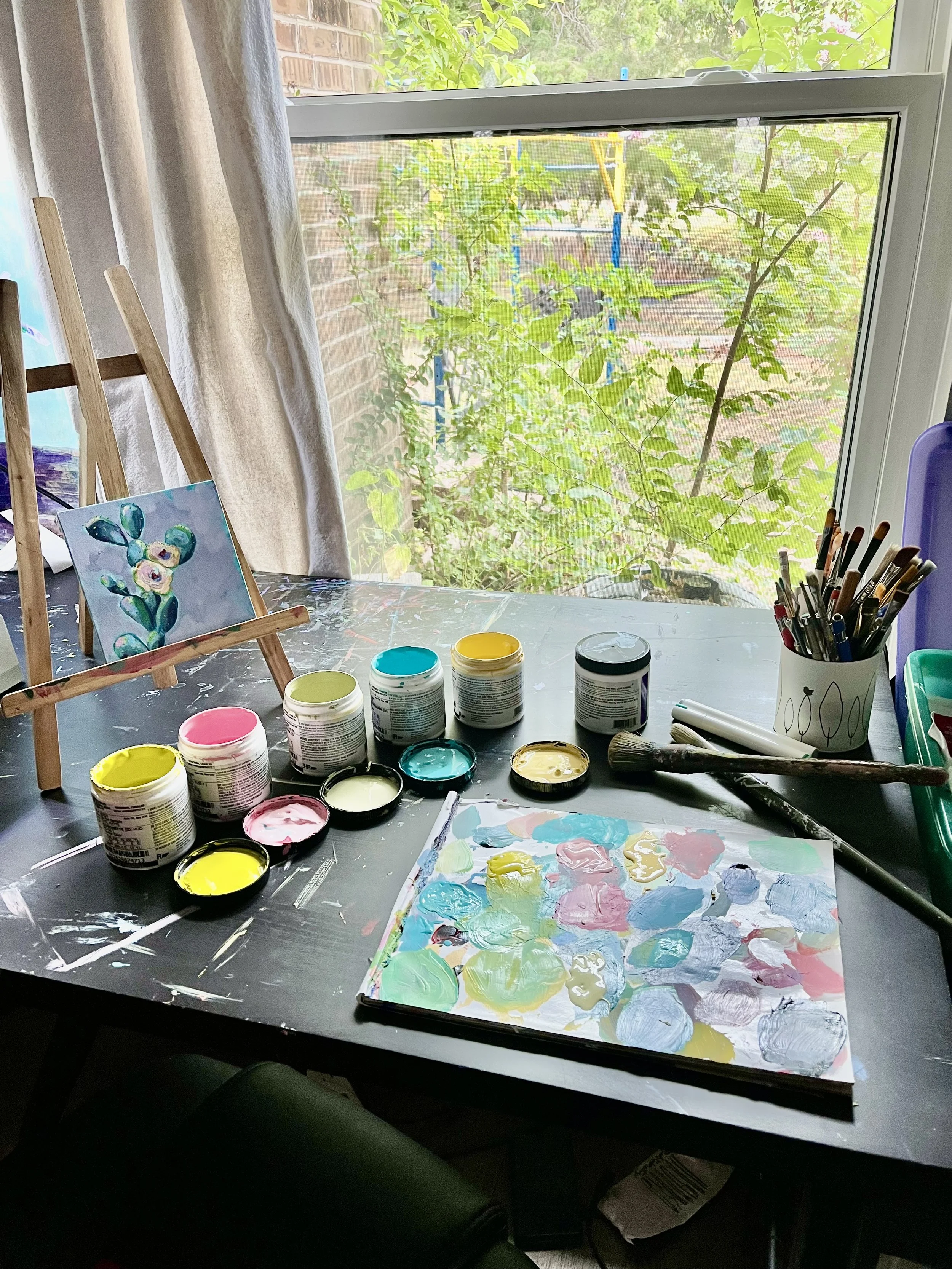 A workspace with a table covered in various acrylic paint jars, brushes, and a palette with mixed paint colors. There is a small easel with a painting of abstract flowers. The background shows a window with green leaves and a brick wall outside.