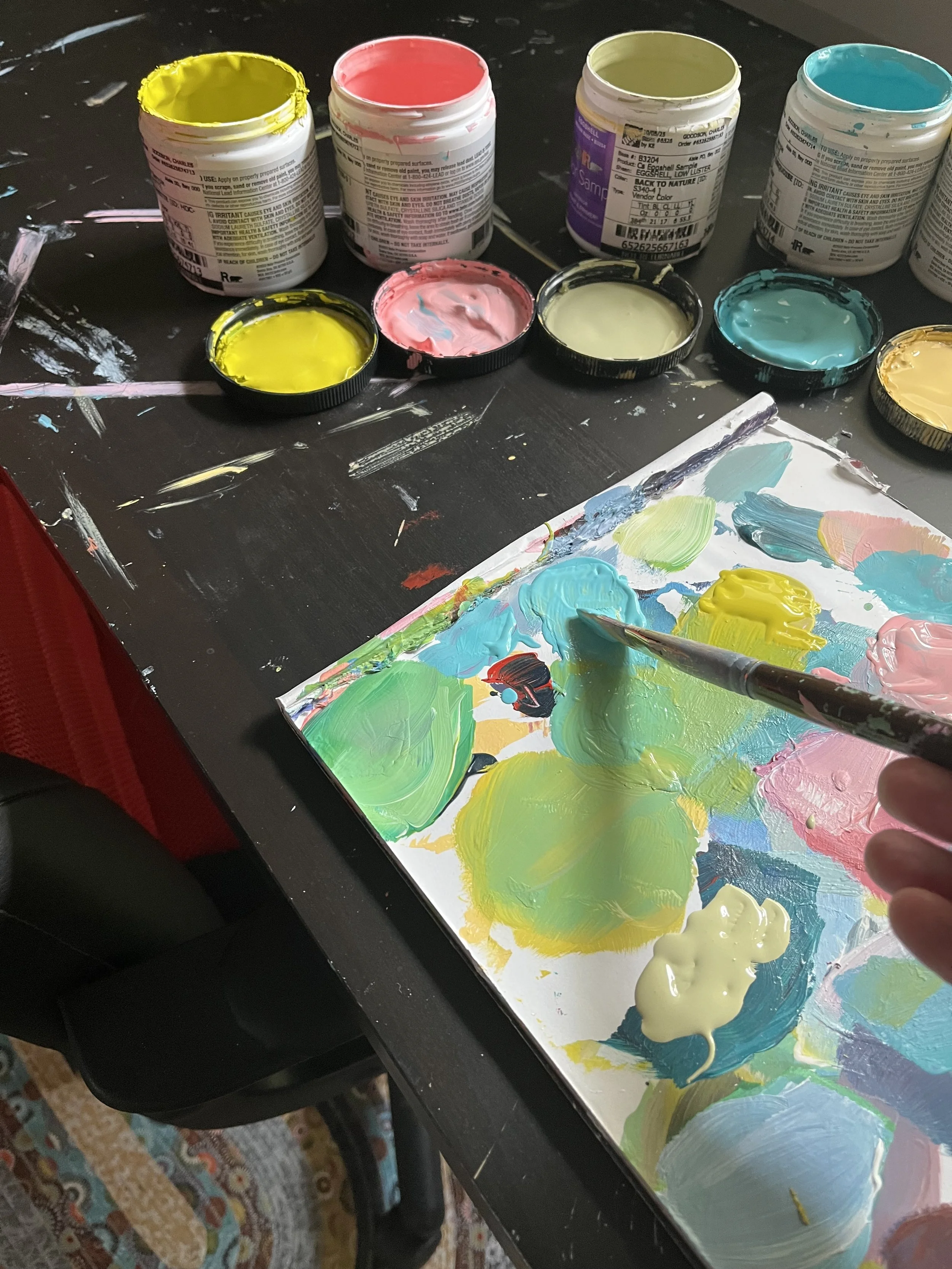 Creating a colorful abstract painting with various shades of yellow, pink, green, blue, and cream on a paper using a paintbrush, with open containers of acrylic paint in different colors on a black surface.