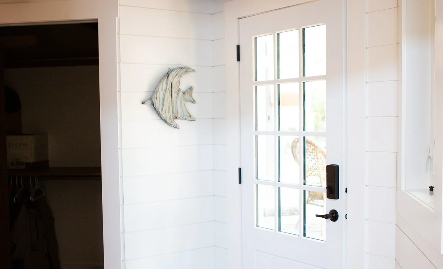 White interior door with windowpanes, leading outside, with a black handle and electronic lock, next to a white wall with a mounted metal fish sculpture. The outside area is partially visible through the door's glass.