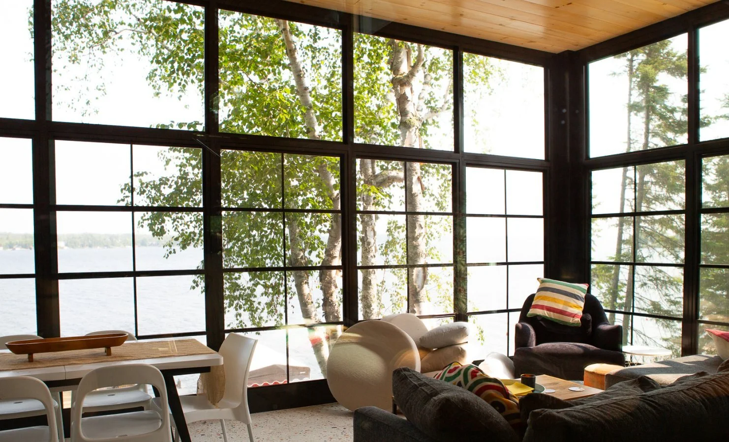 Living room with large windows overlooking trees and water, with dark armchair, white sofa, and a dining table with chairs.