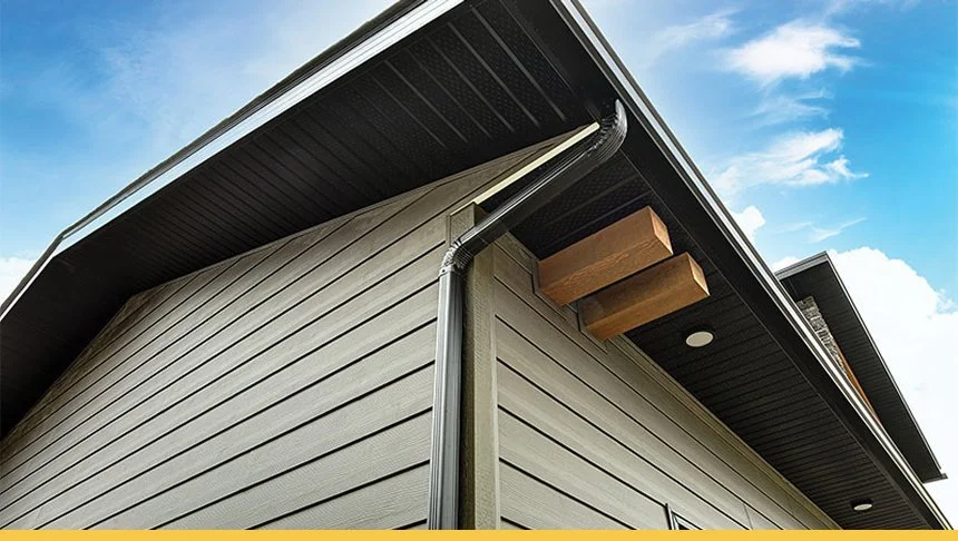 Close-up of the corner of a modern house with gray siding, black roof, and wooden braces under the eaves against a blue sky with clouds.