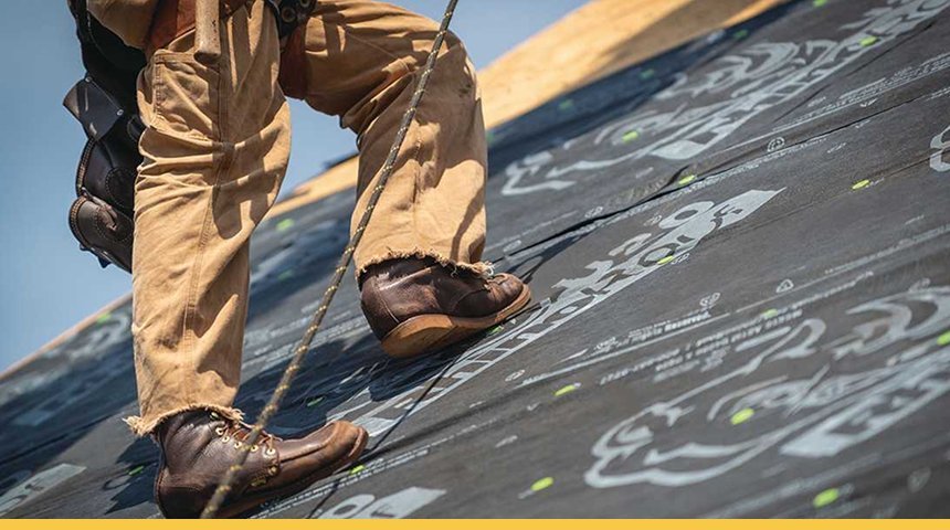 Close-up of a worker's legs and feet on a roofing surface with a black waterproof membrane featuring printed logos, wearing brown work boots, beige pants, and holding a safety rope.