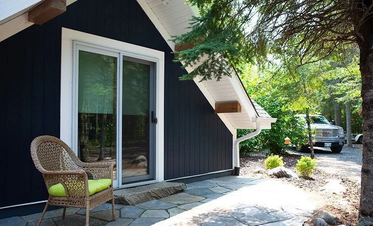 Front porch of a house with a wicker chair with a green cushion, a sliding glass door, and surrounding trees and parked vehicles.