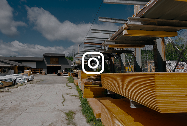 Outdoor scene at a construction or hardware store with stacks of plywood and lumber on racks, a paved driveway, and a building in the background under a partly cloudy sky.