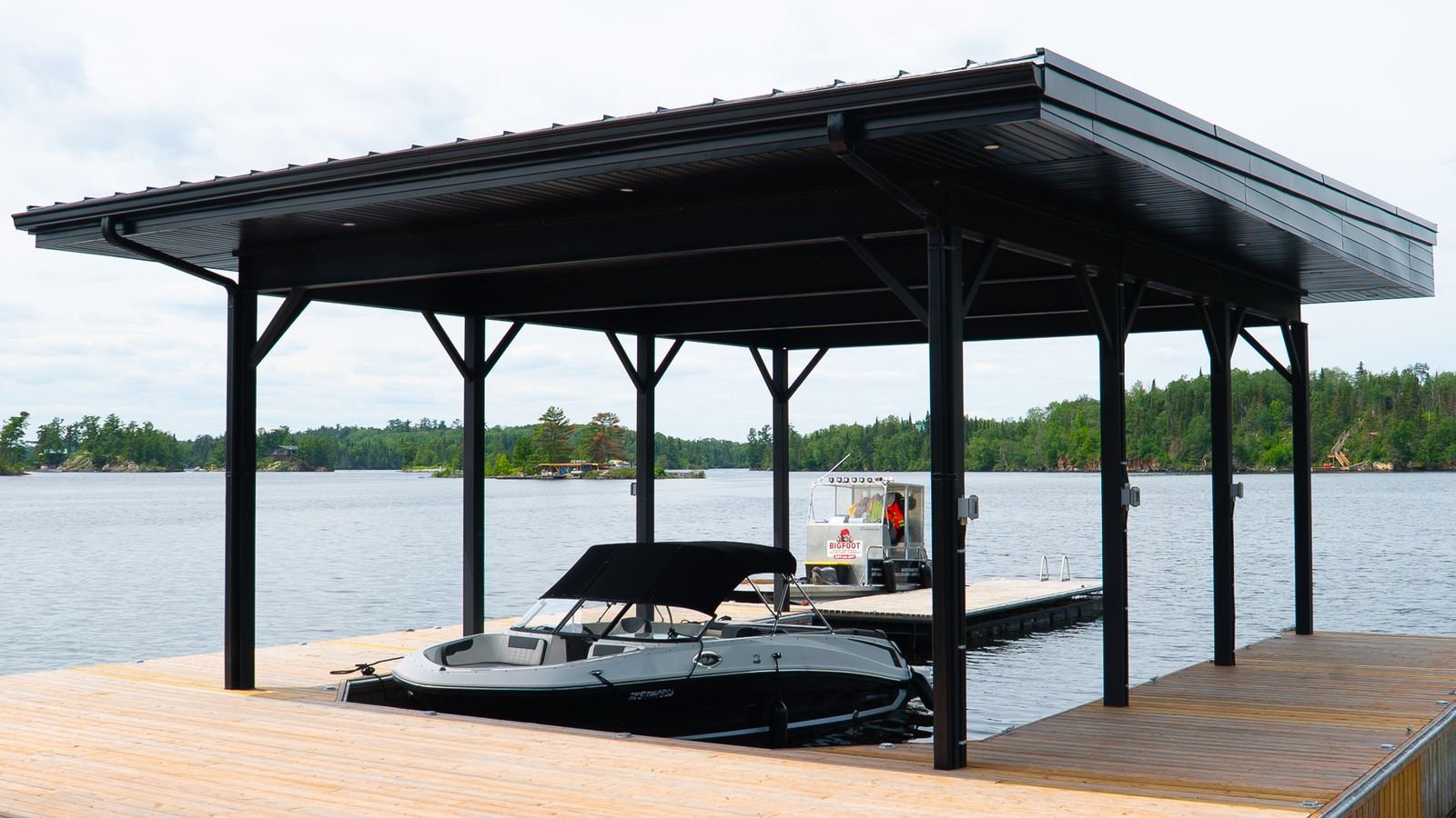 A boat dock with a covered structure over the water, a boat moored at the dock, and a small electric boat nearby, with trees in the background.