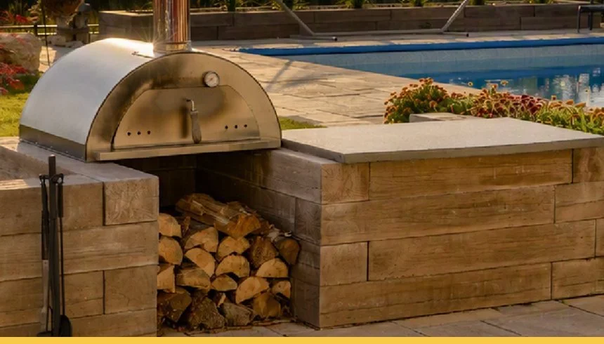 An outdoor wood-fired pizza oven built into a stone and brick structure with firewood stored underneath, near a swimming pool with flowers and patio area in the background.