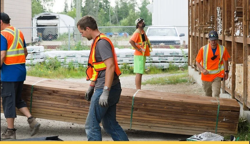 Construction workers in orange safety vests and gloves handling wooden planks on a construction site.
