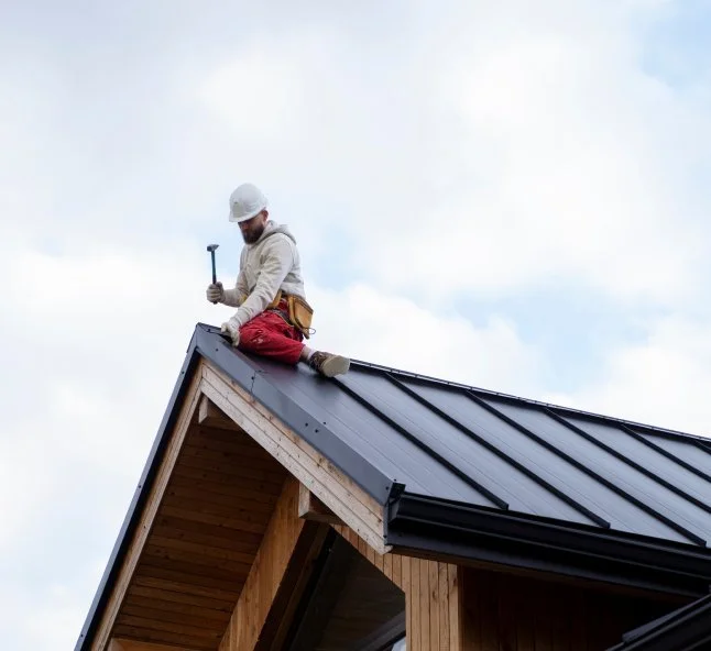 Construction worker on a house roof wearing a white helmet, beige jacket, red pants, and holding a hammer.