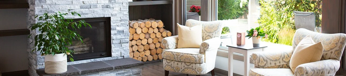 Living room with a stone fireplace, a potted plant, a stack of firewood, two armchairs with patterned upholstery and cushions, a small side table with a flower arrangement and a framed picture, and large windows showing a garden outside.