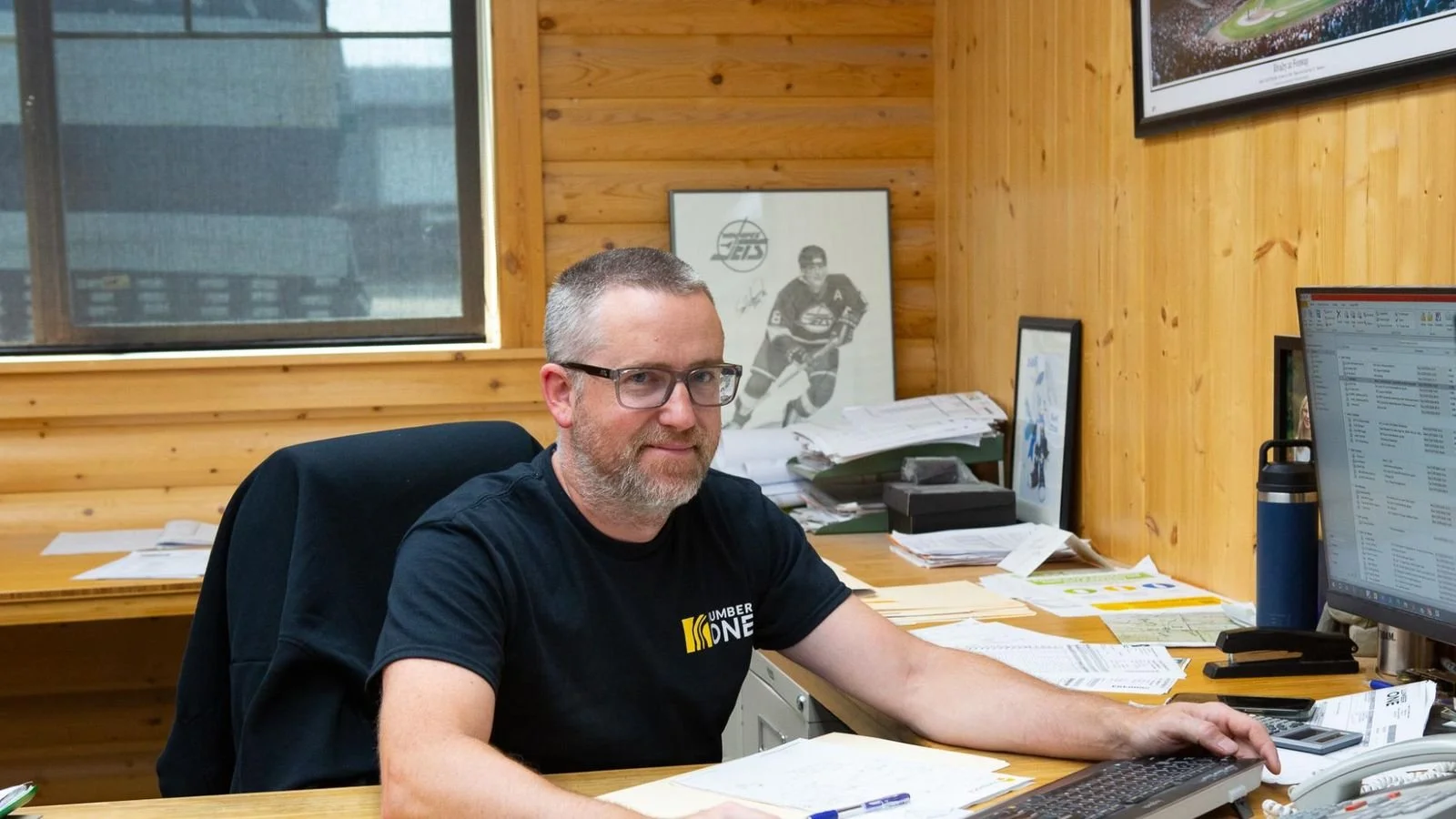 Man with glasses and beard sitting at a cluttered wooden office desk, working on a computer, surrounded by papers, with wood panel walls and framed pictures in the background.