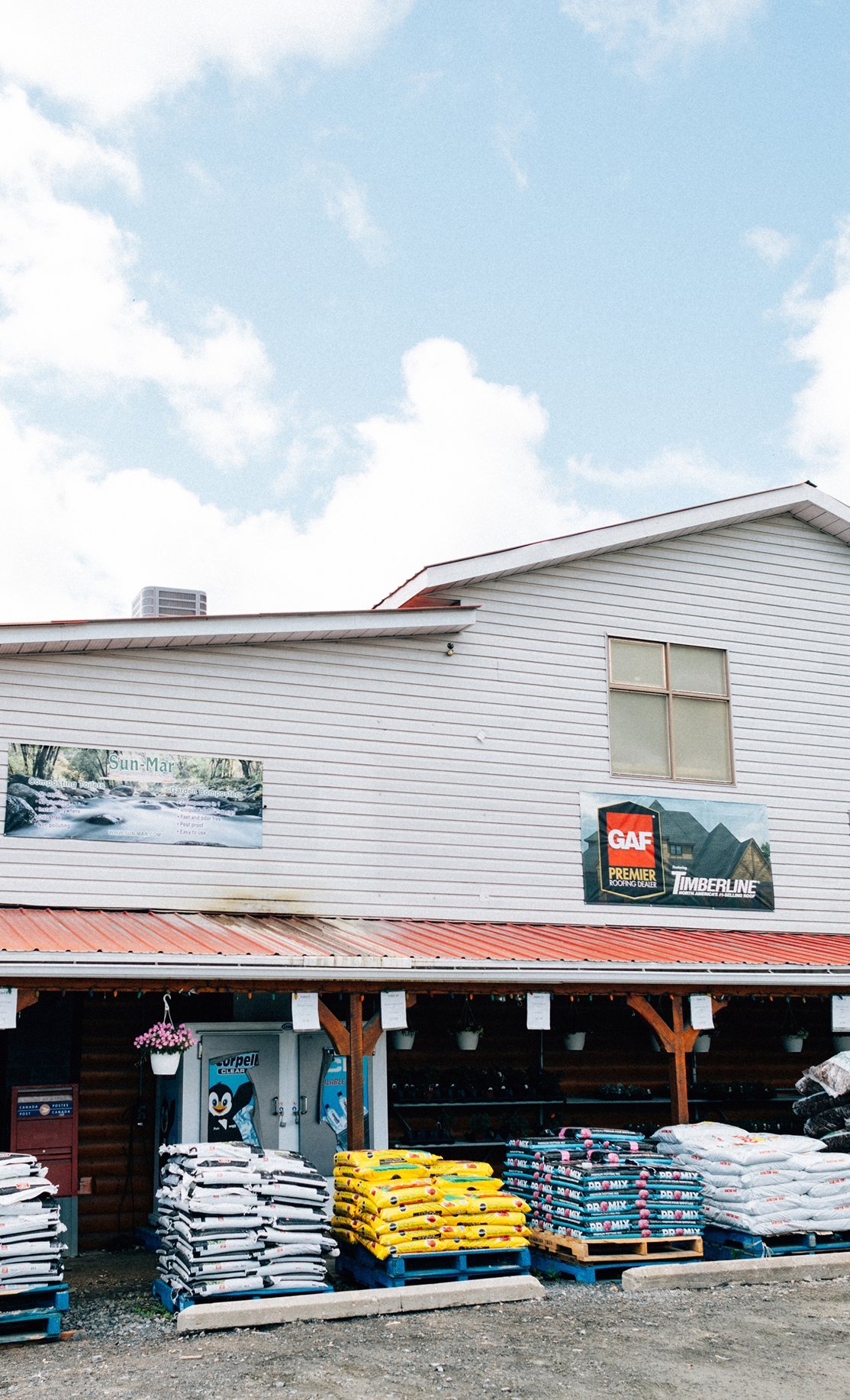 Store with roofing and gardening supplies, stacks of soil bags, and signs for roofing and composting products. The building is white with a partly visible red metal roof, and there are signs advertising roofing services and composting toilets.
