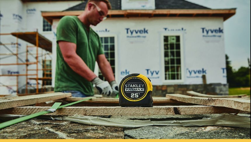 A man working on a construction site with a tape measure in the foreground and a house under construction in the background.