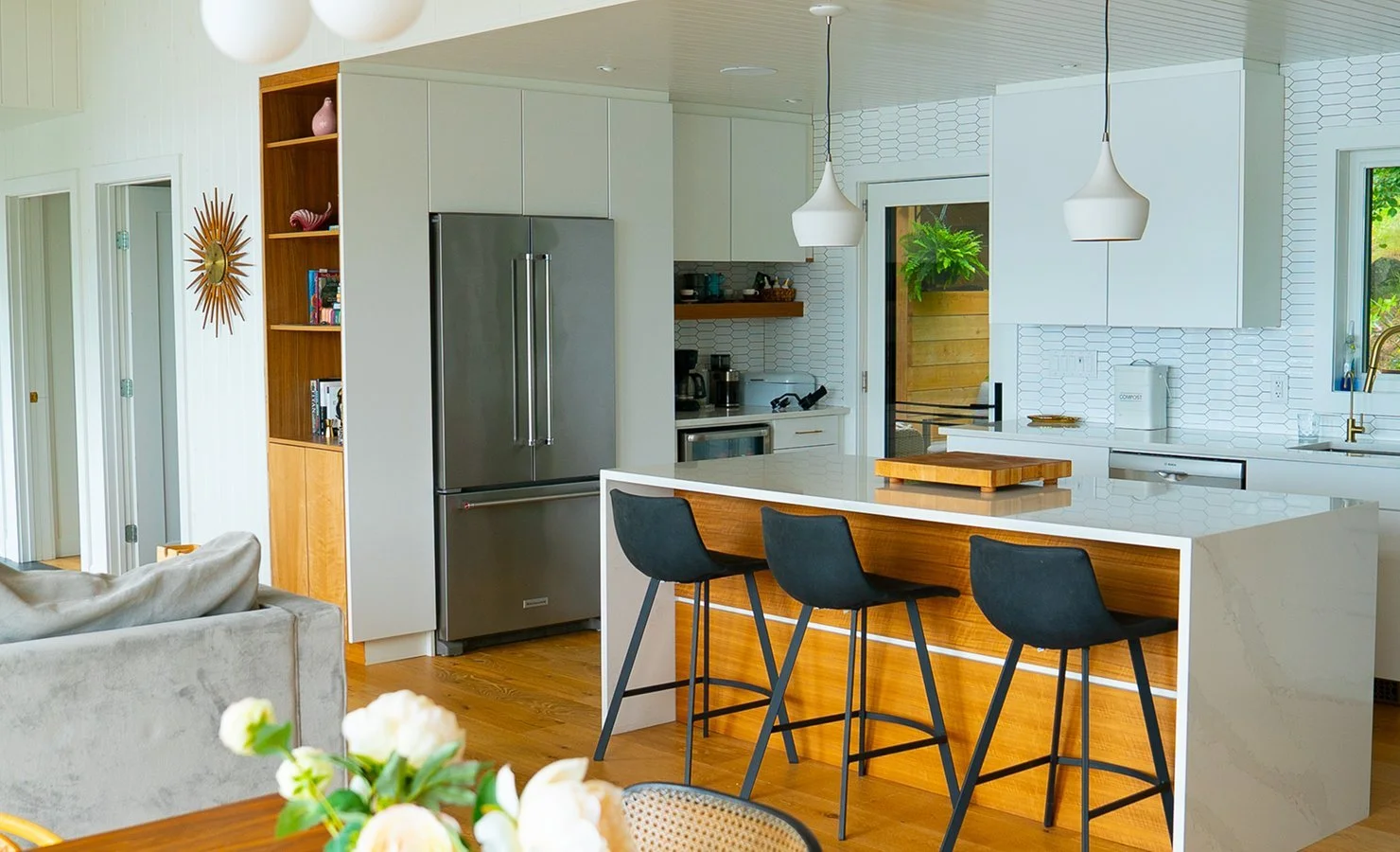 Modern kitchen with white cabinets, a gray refrigerator, a kitchen island with black bar stools, pendant lighting, wood accents, and a sliding glass door