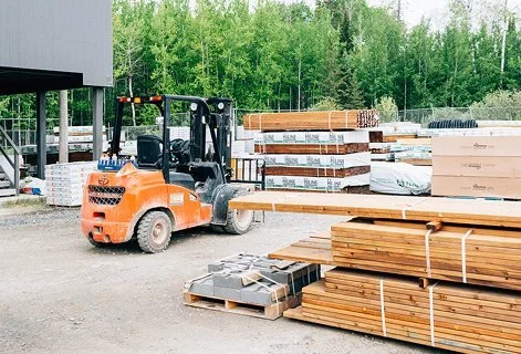 A construction site with a forklift moving wooden planks, with stacks of lumber and building materials around, and trees in the background.