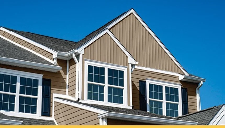 Close-up of a beige house with white trim, multiple windows, and a gabled roof against a blue sky.