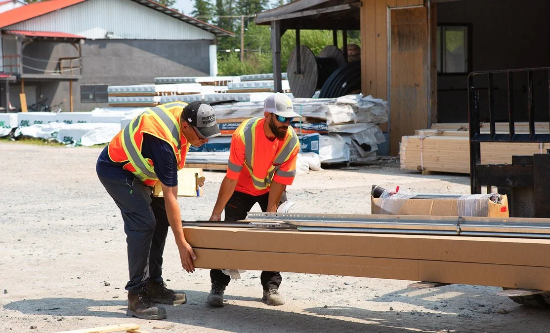Two construction workers wearing safety vests and hats lift a long piece of framing material at a construction site with building materials and equipment in the background.
