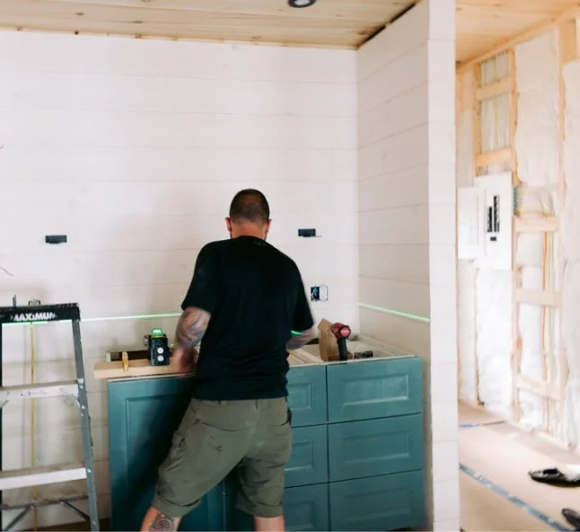 A person with tattoos on their arms working on a kitchen renovation, installing a blue cabinet underneath a white wall, with construction tools nearby.