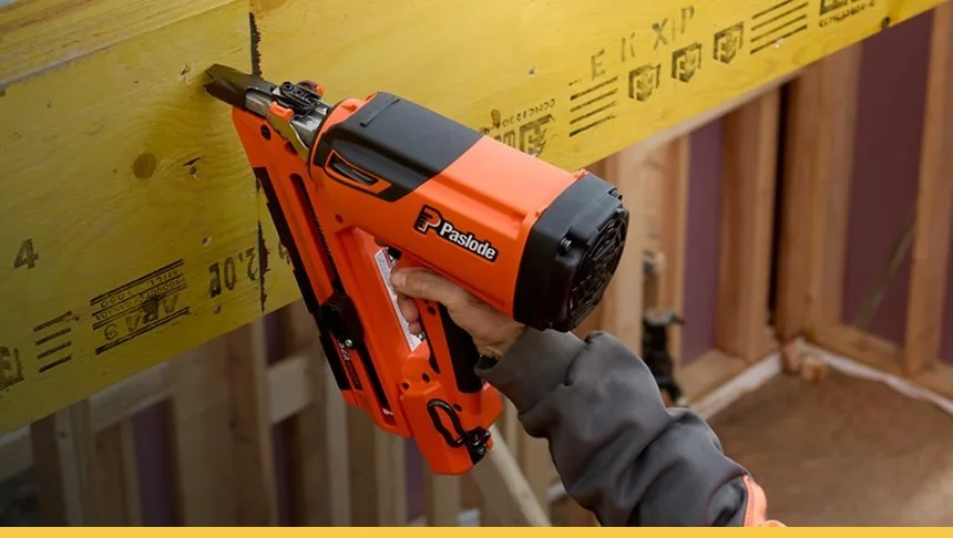 A person using a Paslode cordless nail gun to attach a yellow wooden board to a wall frame during construction.