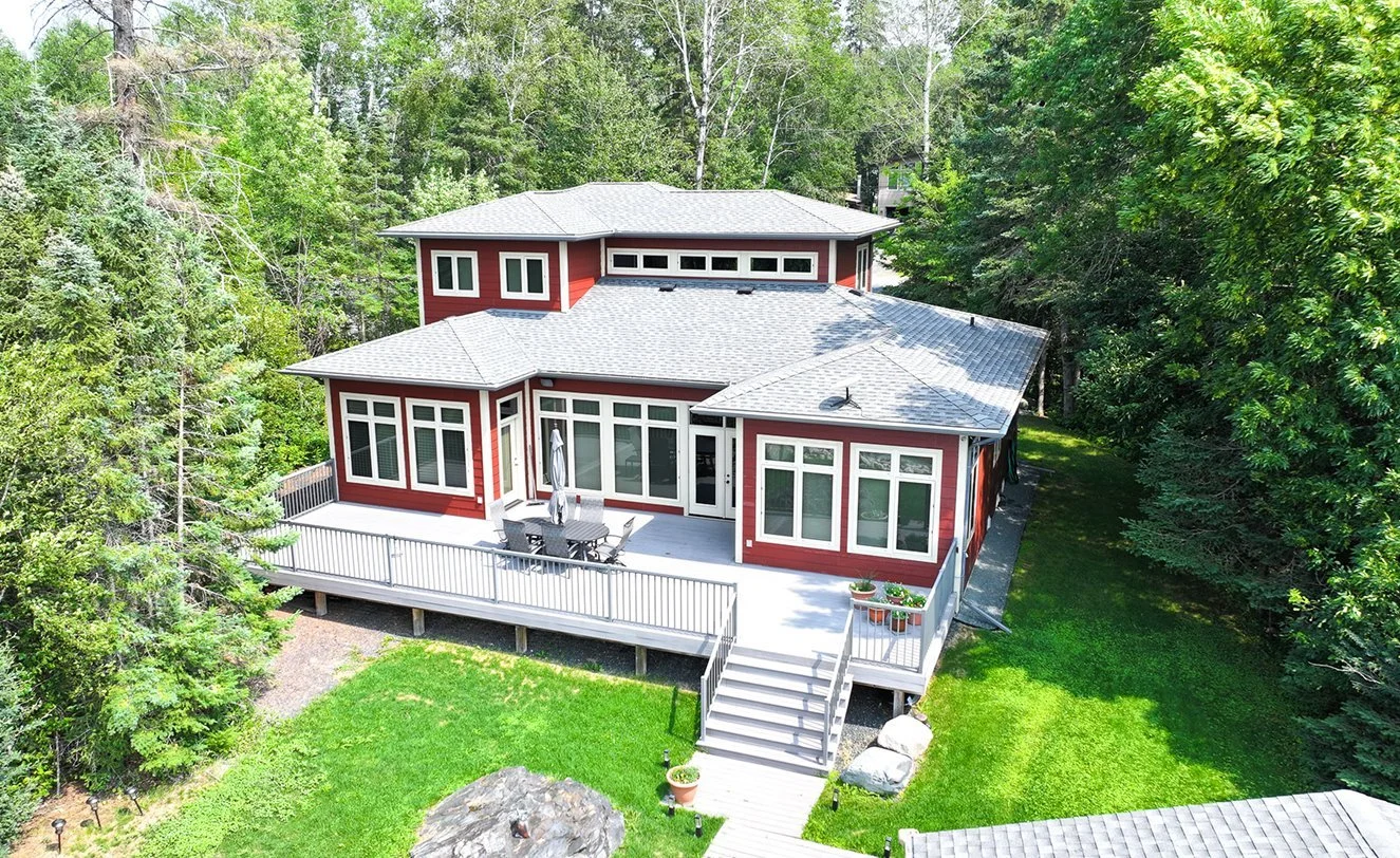 Red house with large windows and a gray roof, surrounded by tall green trees, featuring a wooden deck with outdoor furniture and potted plants.