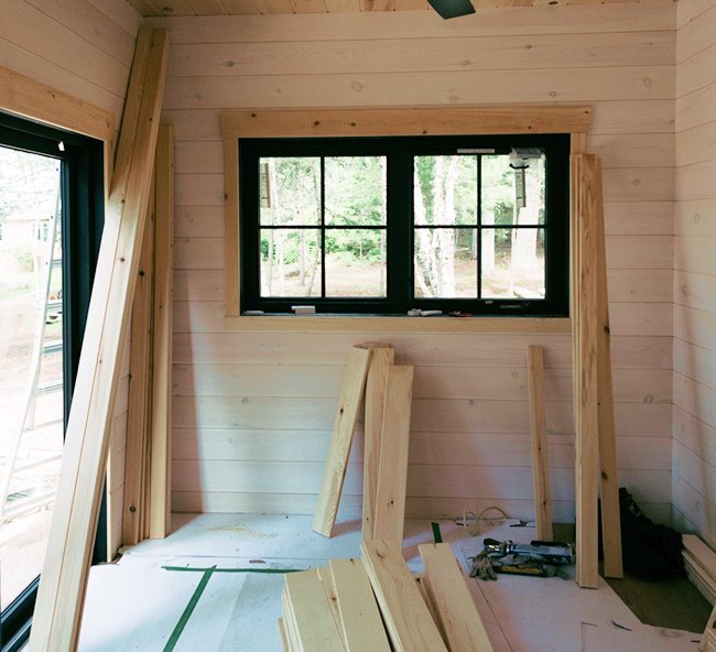 Interior of a room under construction with light-colored wooden walls, a window showing greenery outside, and wooden planks and construction tools inside.