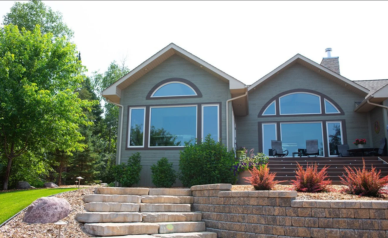 Front view of a modern house with large arched windows, a porch with outdoor seating, and landscaped yard with rocks, bushes, and trees.