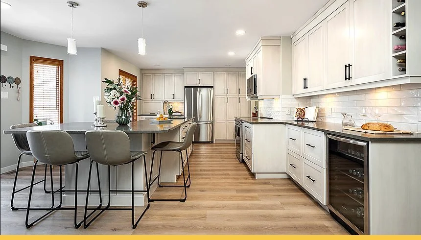 Modern kitchen with white cabinetry, stainless steel appliances, a kitchen island with four gray chairs, wood flooring, and decorative elements like a vase of flowers and a loaf of bread.