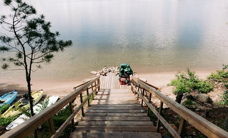 Wooden staircase leading down to a lake, with kayaks on the shore to the left, a small dock, and some greenery and rocks on the right.