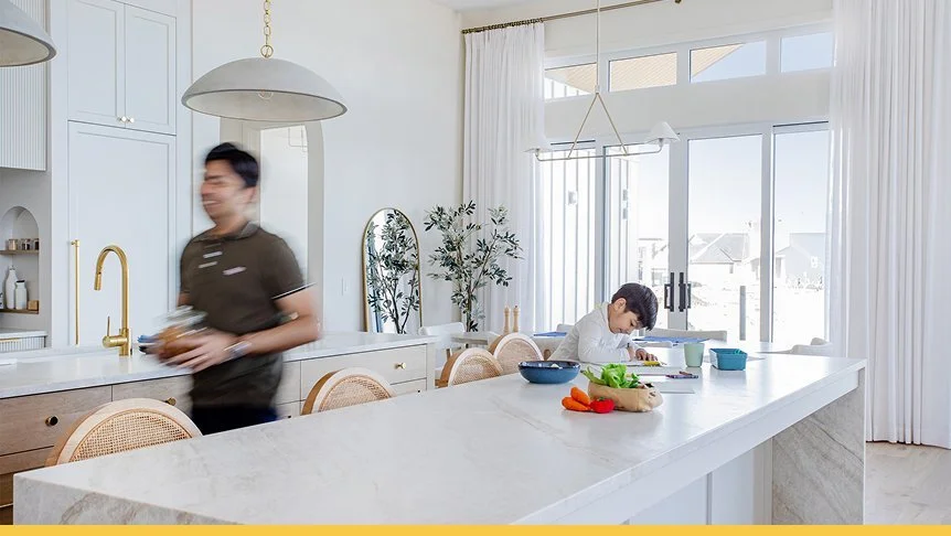 A woman and a boy in a bright, modern kitchen. The woman is walking past the kitchen island with a blurred motion, holding a bowl. The boy is sitting at the kitchen island, focused on preparing food with vegetables and kitchenware on the counter. Large windows with white curtains let in natural light.