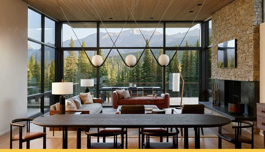 Modern living room with large floor-to-ceiling windows showcasing mountain and forest views, featuring a stone fireplace, contemporary lighting, and stylish furniture.