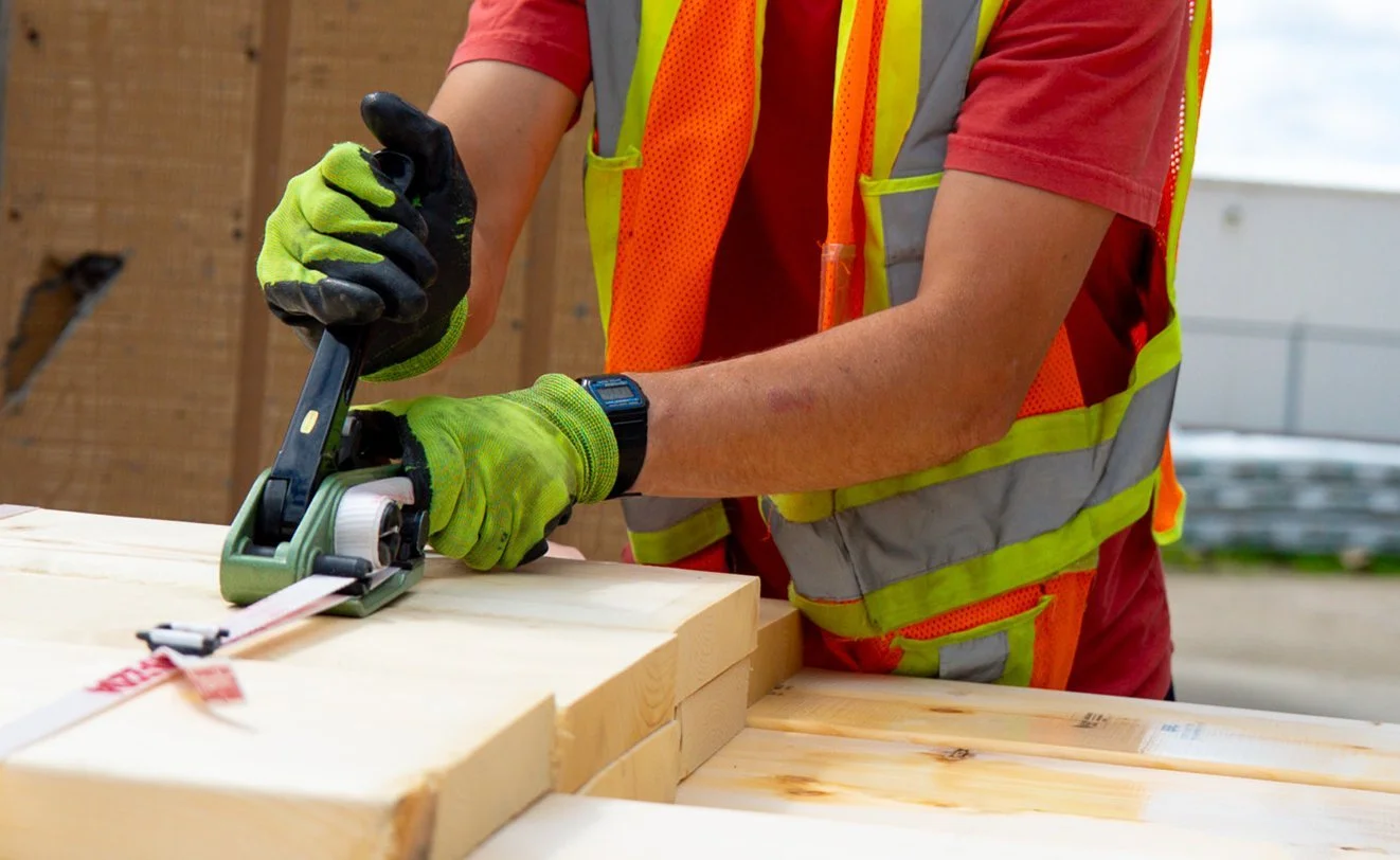 A construction worker wearing a safety vest and gloves using a power planer on a piece of wood outdoors.