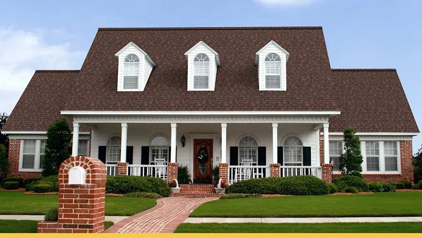 Large two-story house with a brick and white exterior, a steep roof with three dormer windows, a front porch with white columns, a brick walkway, and a well-maintained green lawn with shrubs.