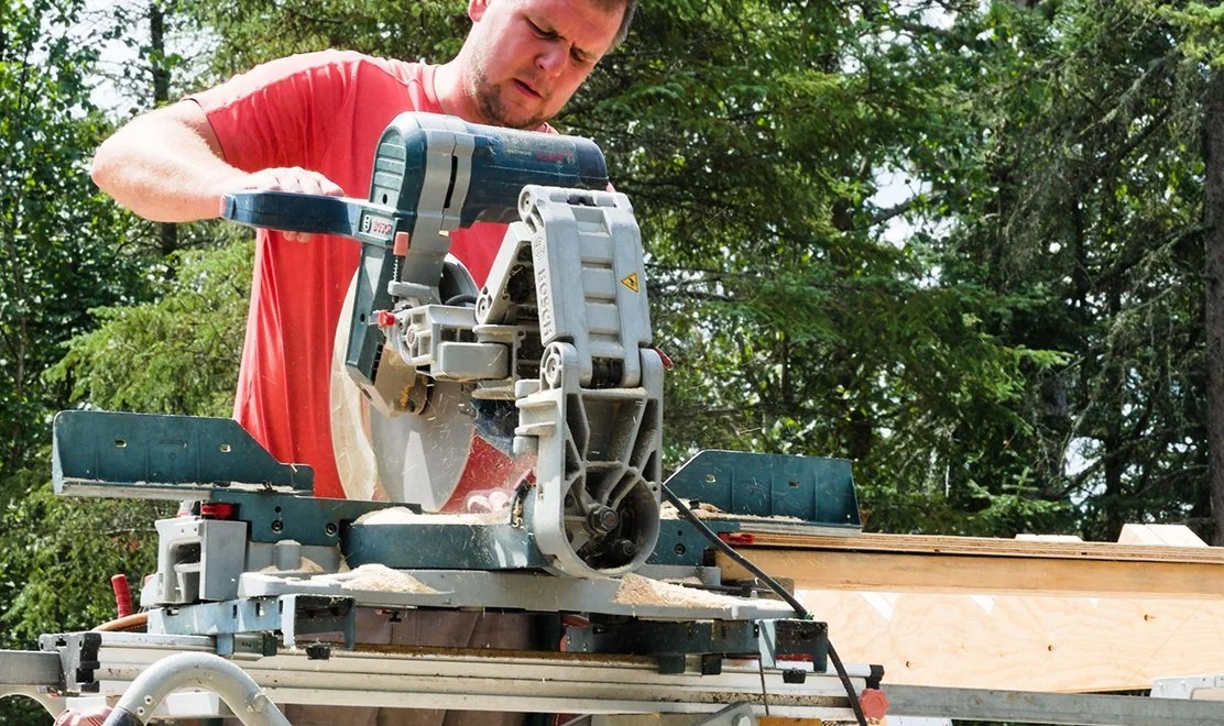 A man in a red t-shirt using a sliding miter saw outdoors with trees in the background.