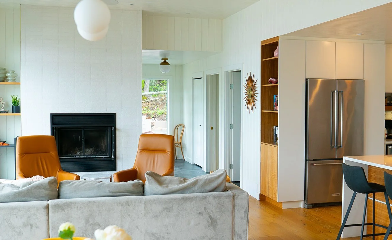 Living room with beige sofa, two orange armchairs, a white brick fireplace, wooden bookshelf, stainless steel refrigerator, and a kitchen counter with bar chairs.