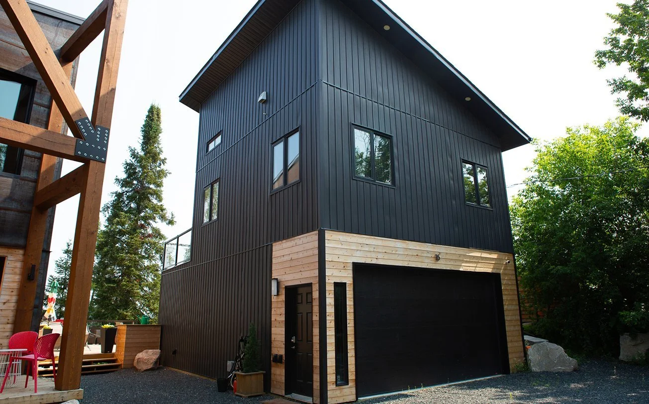 A modern three-story house with black vertical siding and a wood-paneled garage entrance, surrounded by trees and neighboring structures.