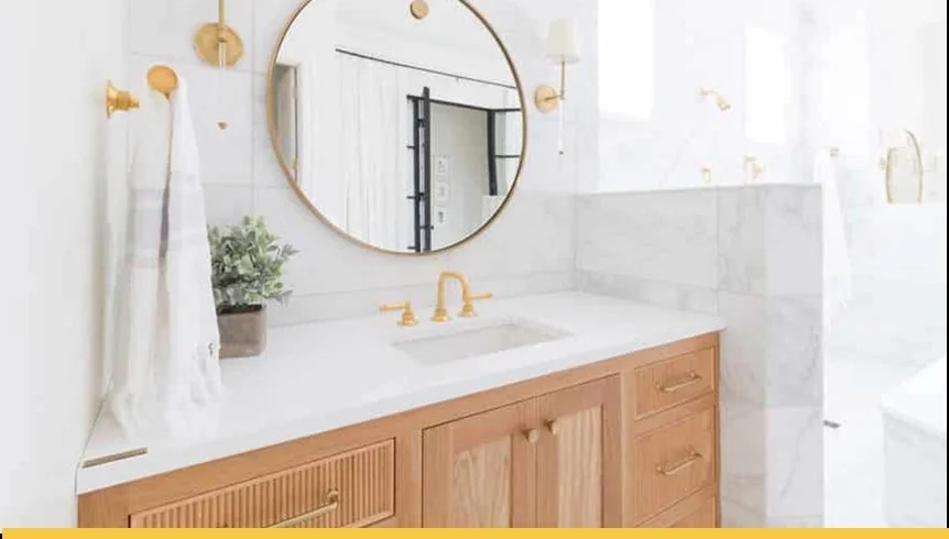Bathroom vanity with a wooden cabinet, white countertop, gold fixtures, a round mirror, and a towel with a plant in a pot beside the sink.