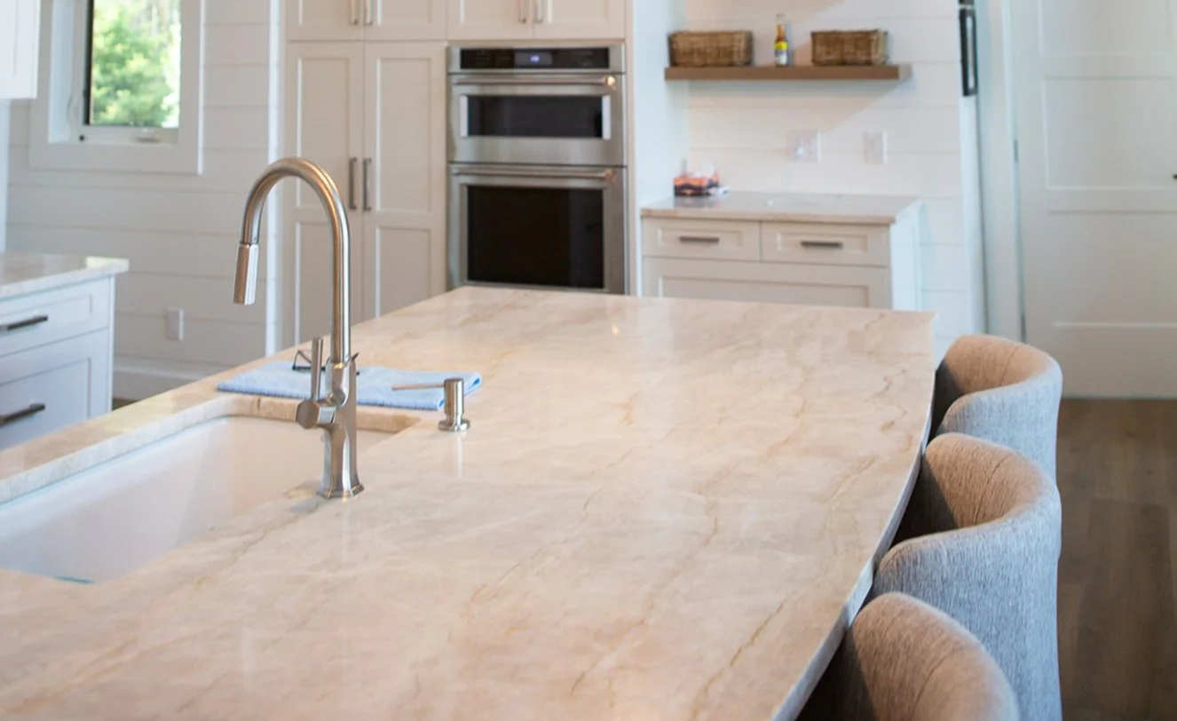 Modern kitchen with a marble countertop island, stainless steel faucet, cream-colored chairs, built-in oven, and upper shelves with baskets and bottles.