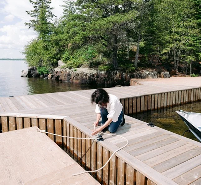 A woman kneeling on a wooden dock by the water, tying or securing rope to the dock's edge, with trees and rocks in the background.