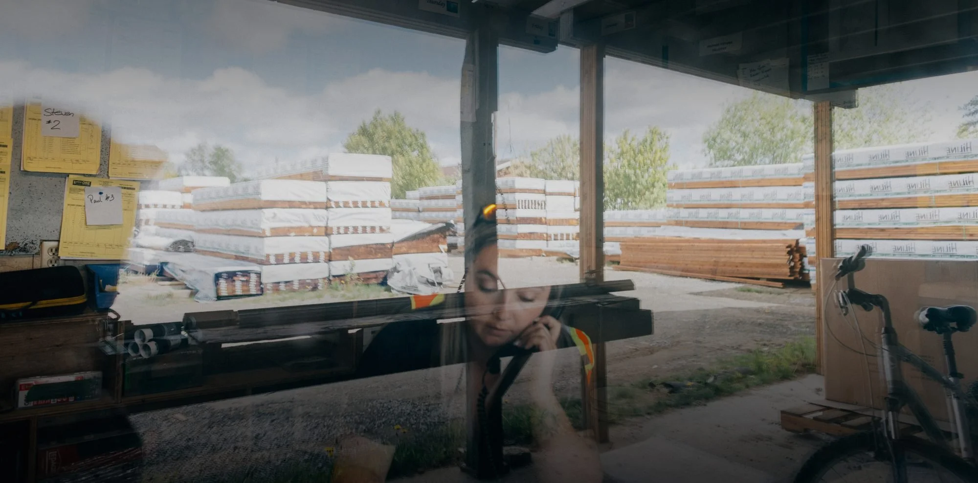 A reflection of a woman in an indoor space with construction materials outside including stacked lumber wrapped in plastic and a stack of wooden planks, a person's face is partially visible in the reflection.